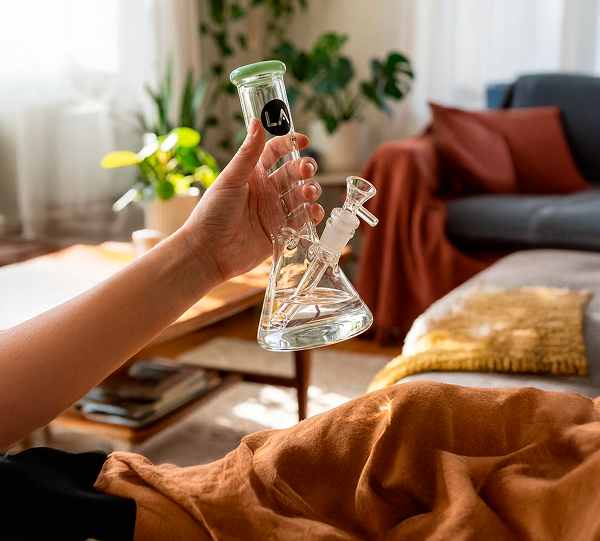Handheld, angled view of the LA Pipes Color Accented Mini Beaker Bong in black-on‑black glass with a green-rimmed mouthpiece, shown in a sunlit living room.