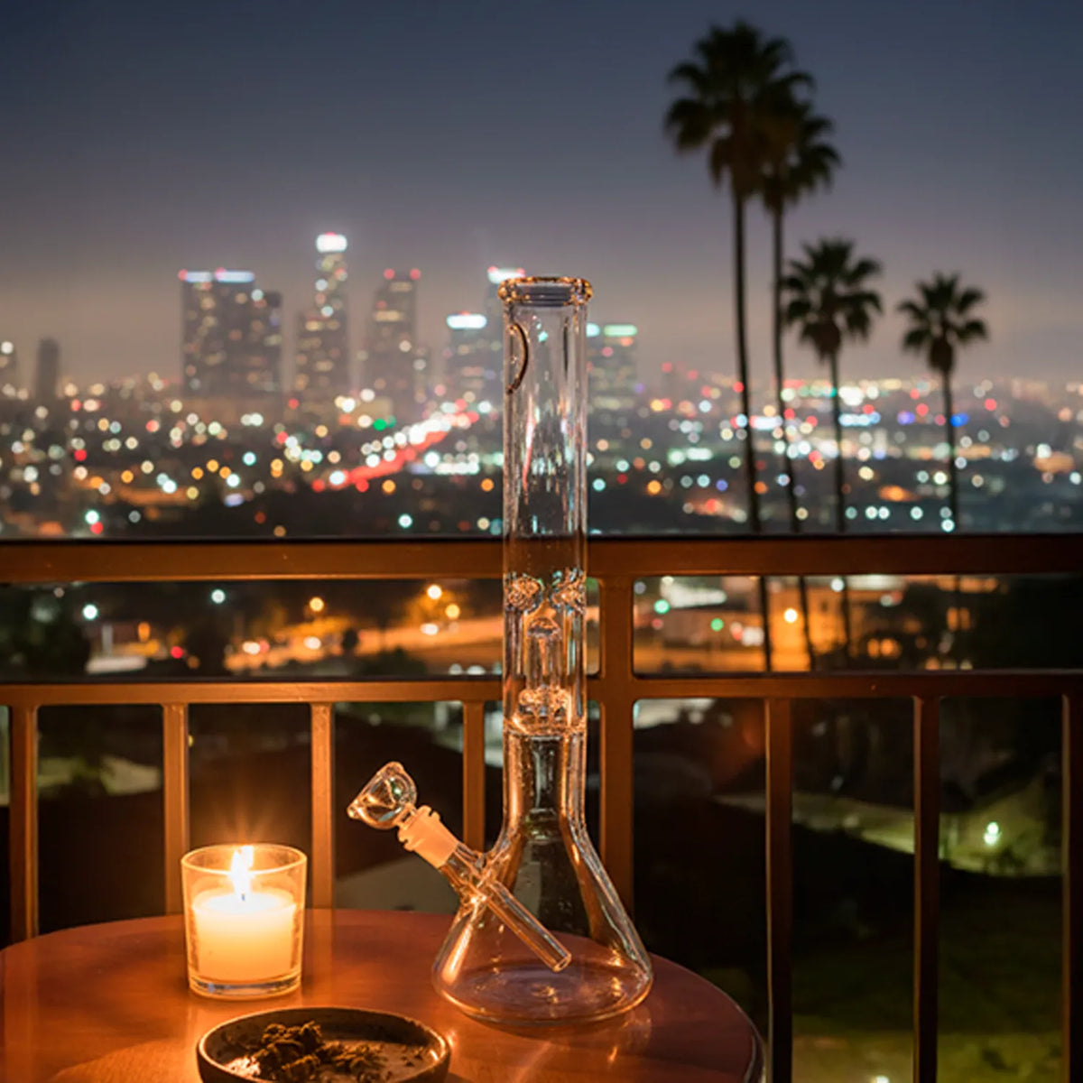 Evening view of the clear 16" LA Pipes 16" Clear UFO Showerhead Perc Beaker Bong on a balcony table with city lights and palm trees blurred behind.