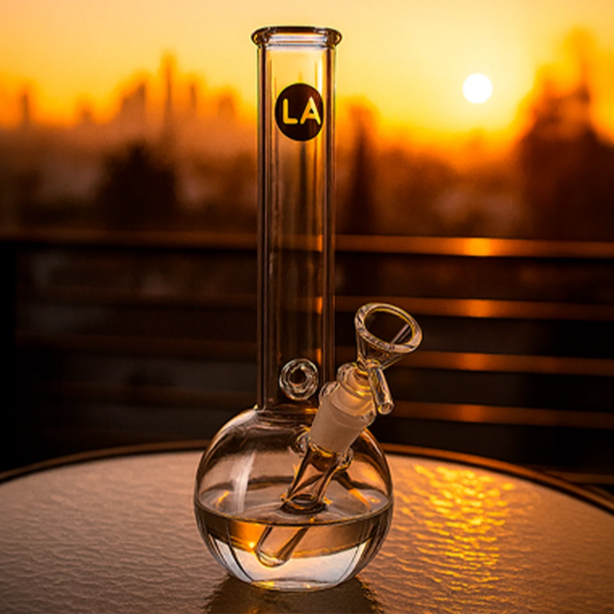 Sunset-lit close-up of the LA Pipes Baller Bubble Base Bong centered on a table, the clear glass sphere base and straight neck reflecting warm orange light.